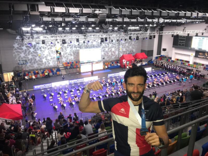 Hombre celebrando con una medalla en un evento deportivo en una arena grande con público y otros atletas en el fondo.