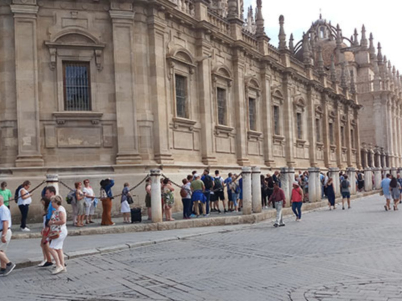 Una calle con gente caminando frente a un edificio monumental de piedra, posiblemente una catedral o iglesia histórica. La arquitectura es clásica con detalles ornamentales y columnas, y el cielo está despejado. La escena parece ser en un día soleado, con la gente vestida de manera casual para el clima.