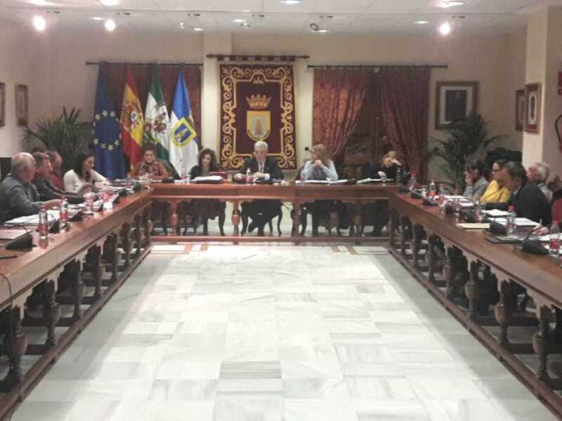 Sala de reuniones con mesas largas, sillas y personas sentadas. Banderas europeas y estatales en el fondo. Tapices y cuadros decorativos.