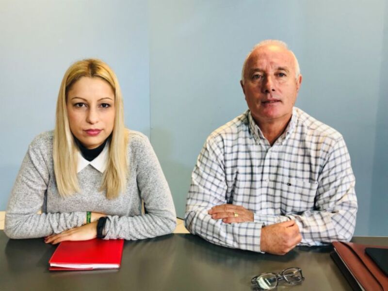 Dos personas sentadas en una mesa, uno con camisa a cuadros y el otro con suéter gris. En la mesa hay un bloc de notas rojo, gafas y una pluma. El fondo es azul claro.