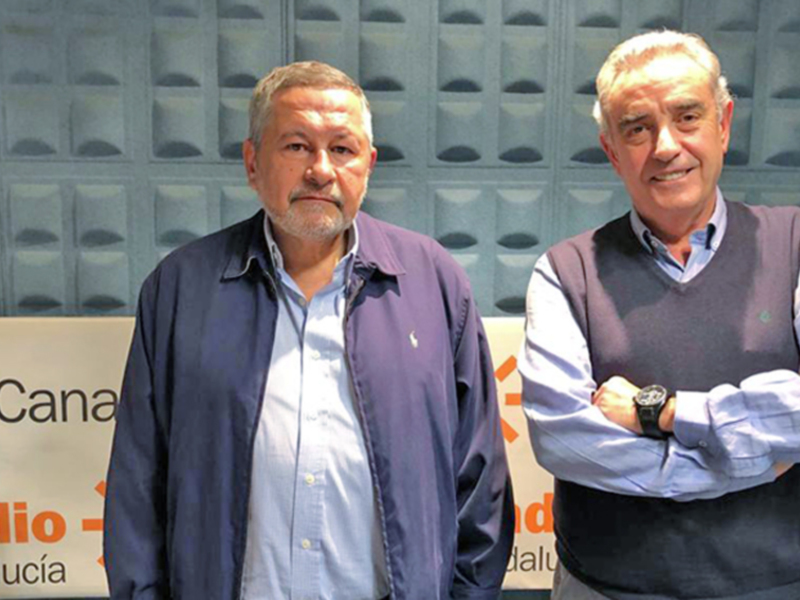Dos hombres posan frente a un fondo con la marca de radio 'Cana Radio'.