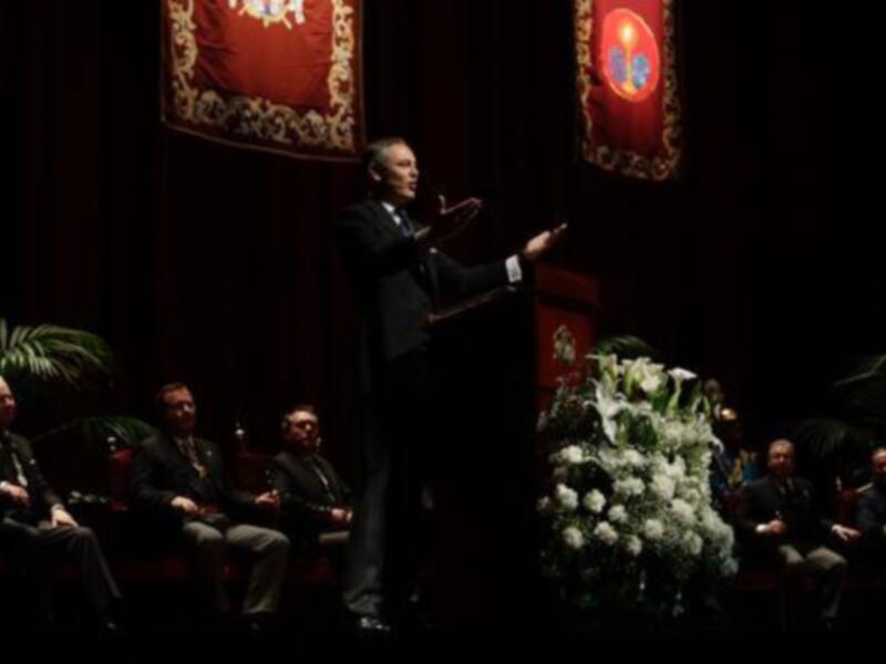 Una persona hablando en un escenario con una bandera y flores blancas detrás.
