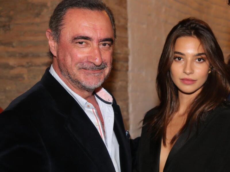 A man and a woman standing together, both dressed in formal attire. The man is wearing a dark suit jacket over a white shirt with a red and white striped tie, while the woman is in a black dress. They are standing in front of a brick wall.
