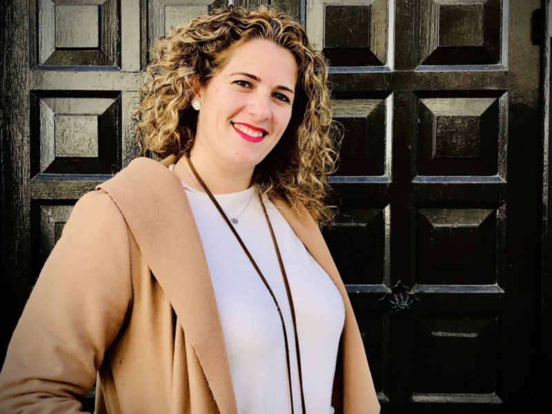 A una mujer con cabello rizado y una sonrisa amigable. Lleva un abrigo beige y una camiseta blanca, con un collar de cuentas. La背景 es una puerta de madera oscura con patrones cuadrados.