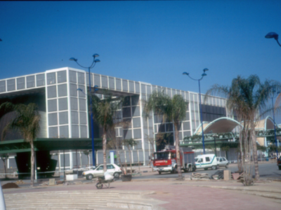 Edificio moderno con estructura de vidrio y metal, ubicado en una zona con árboles y vehículos a la entrada. El cielo es azul claro y el entorno parece ser un lugar urbano o industrial.