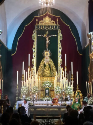 Imagen de una iglesia con un altar dorado y una imagen central. En el fondo, hay una cruz con un crucificado y una figura dorada en la parte superior. La imagen está iluminada por las velas encendidas y una lámpara de araña. La decoración es elegante con detalles dorados y un fondo rojo.