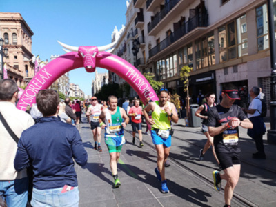 Corredores en una carrera urbana, con una estructura inflable rosa con cuernos en el camino.