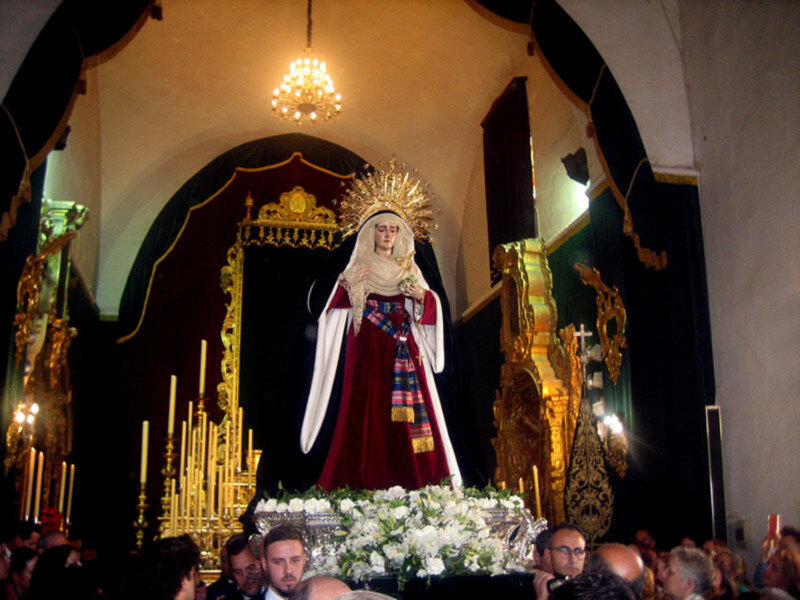 Procesión religiosa con imagen de la Virgen María en un templo, rodeada de fieles y adornada con flores y candelabros dorados.