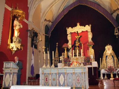 Imagen de una iglesia con un altar decorado y una estatua en el后台. Un hombre se encuentra frente a un púlpito, posiblemente dando una oración o discursando. La decoración incluye flores y elementos religiosos.