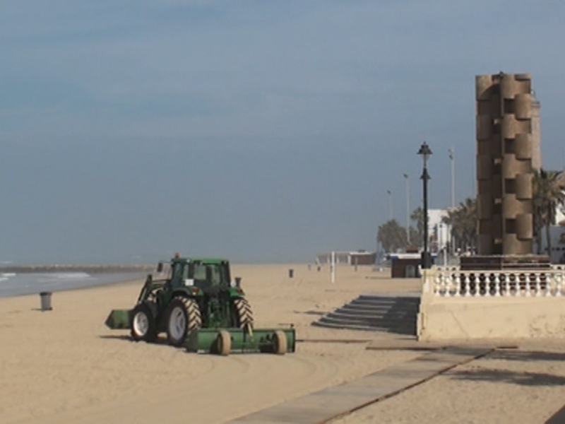Una máquina agrícola verde está trabajando en la arena de una playa. Al fondo, se pueden ver edificios y un monumento con forma de torre. El cielo es azul claro, indicando buen tiempo.