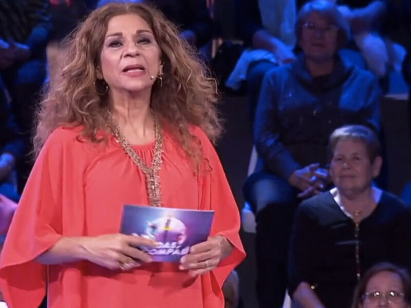 Una mujer con una camisa roja y una cadena dorada, sosteniendo un folleto mientras habla frente a un público en un evento formal.