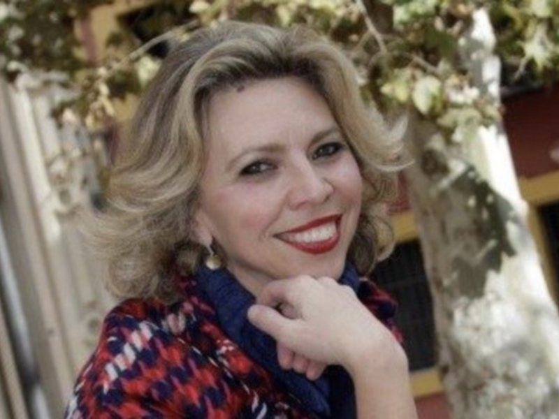 Una mujer con cabello largo y ondulado, vistiendo una chaqueta de cuadros rojos y azules. La imagen muestra detalles del entorno, como una ventana con marco de madera y un árbol en el fondo.