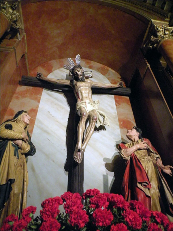Cristo crucificado en una iglesia, rodeado de flores rojas. Estatuas religiosas en el fondo.