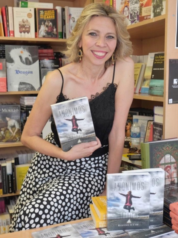 A una mujer sonriente sosteniendo un libro titulado "Anónimos" en una librería. El fondo incluye estanterías repletas de libros variados, destacando una colección de novelas con portadas en diferentes colores y títulos.