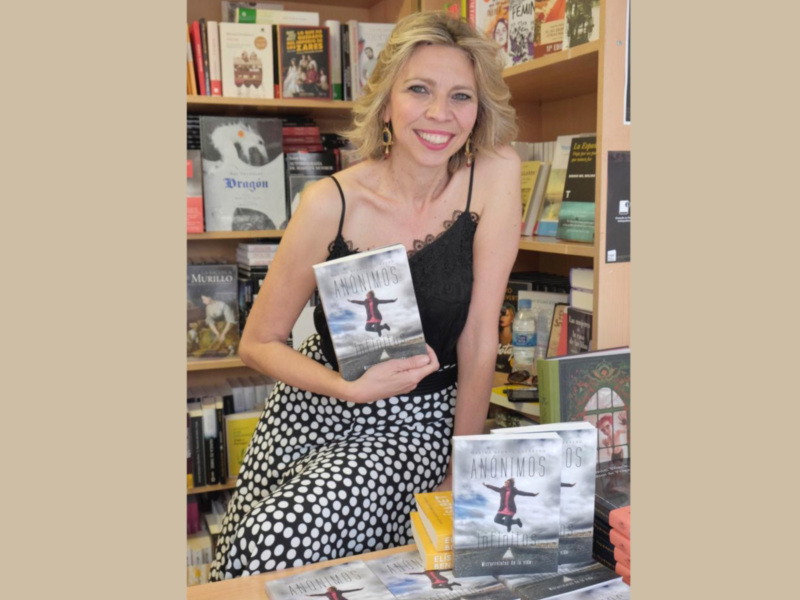A una mujer sonriente sosteniendo un libro en una librería llena de libros. La estantería está repleta de títulos variados, destacando una selección de novelas y otros géneros. La mujer está vestida con un vestido negro a lunares blancos y una camiseta negra. La escena transmite un ambiente de lectura y conocimiento.