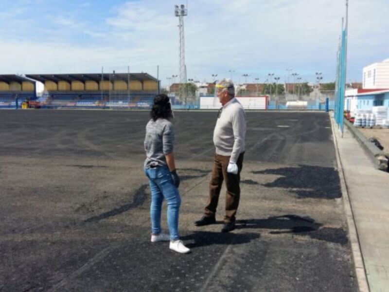 Dos personas caminan por una pista de atletismo vacía, con estructuras de luz en el fondo y edificios al lado.