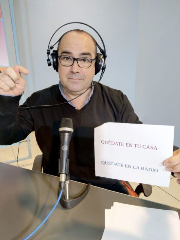 Un hombre con auriculares y una camiseta negra sostiene un papel blanco que dice "QUÉDATE EN LA RADIO". Está en frente de un micrófono y una pantalla.
