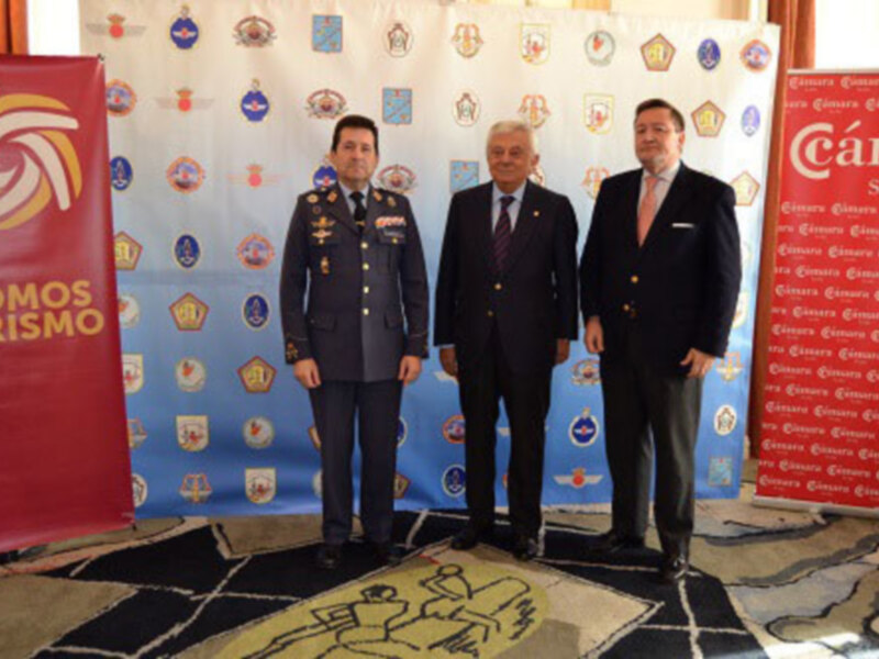 Tres personas posan frente a un fondo con logos de empresas turísticas, uno de ellos con uniforme militar, en un evento formal.