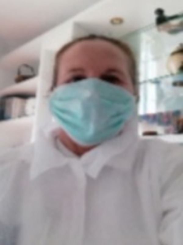 Persona con camisa blanca y mascarilla médica, posando en un entorno interno con estantes en el fondo.