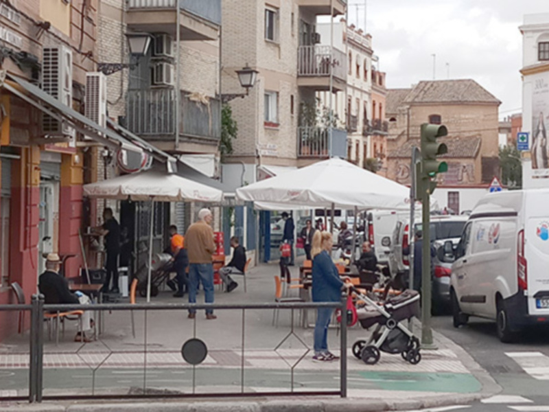 Un bar con mesas y sillas al aire libre, personas sentadas y caminando, edificios al fondo y un carril de tráfico con una señal de semáforo verde.