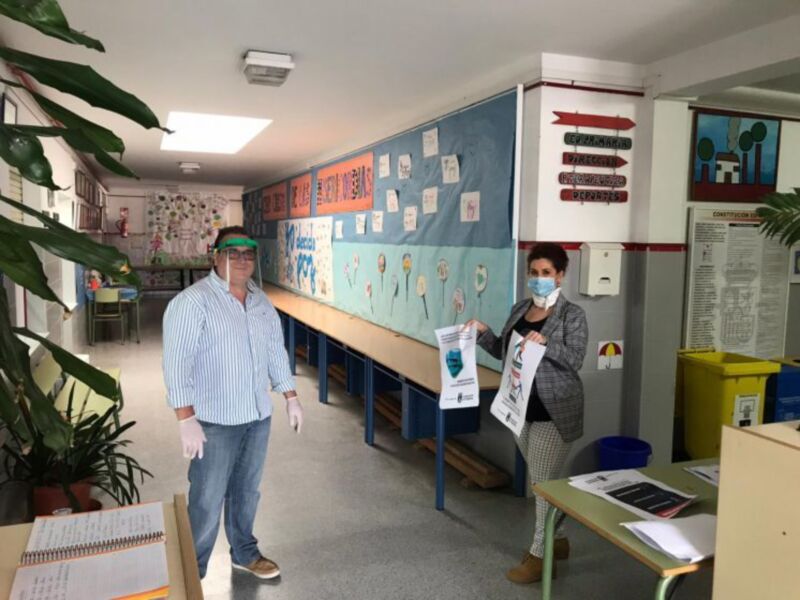 Un hombre y una mujer en un aula, ambos con mascarillas y guantes, frente a un pizarrón con mensajes de seguridad y un cartel informativo. La sala está decorada con plantas y carteles educativos.