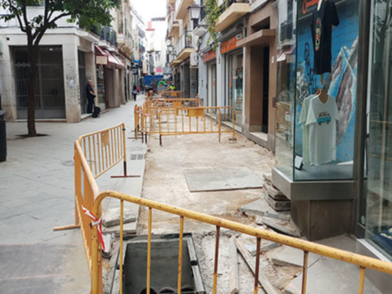 Una calle urbana con edificios de varios pisos y comercios. Hay un poste de teléfono antiguo a la izquierda, barreras amarillas en el centro para indicar obras de construcción y un escaparate con una camiseta colgada en la derecha. La calle parece estar en proceso de renovación o mantenimiento.