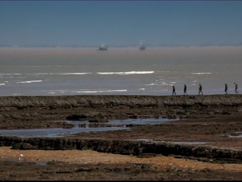Personas caminando por la costa rocosa junto al mar, con barcos en el horizonte.