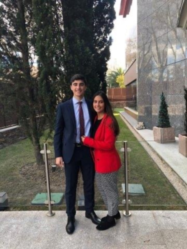 Dos personas posando en la entrada de un edificio moderno, con árboles y una escena urbana al fondo.
