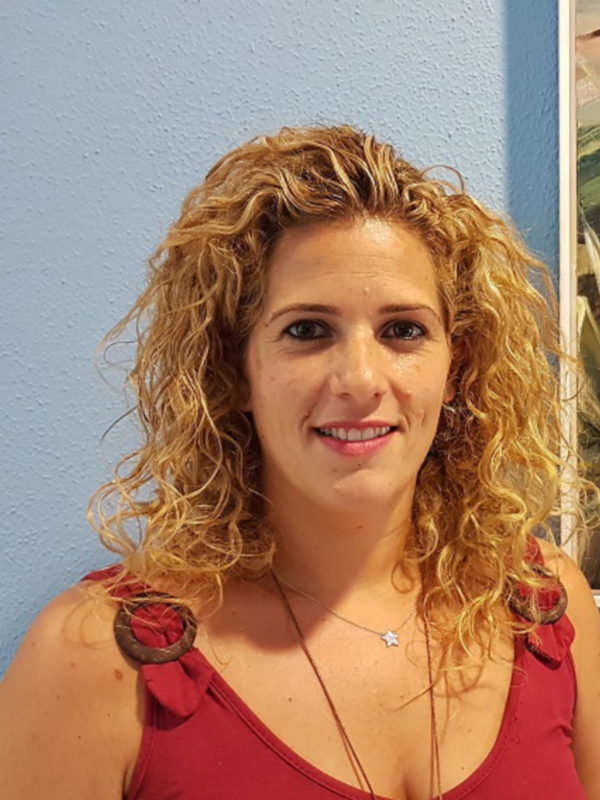 Una mujer con cabello rizado rubio, vistiendo un vestido rojo y una joya en el hombro. Fondo azul claro con un cuadro de paisaje al lado.
