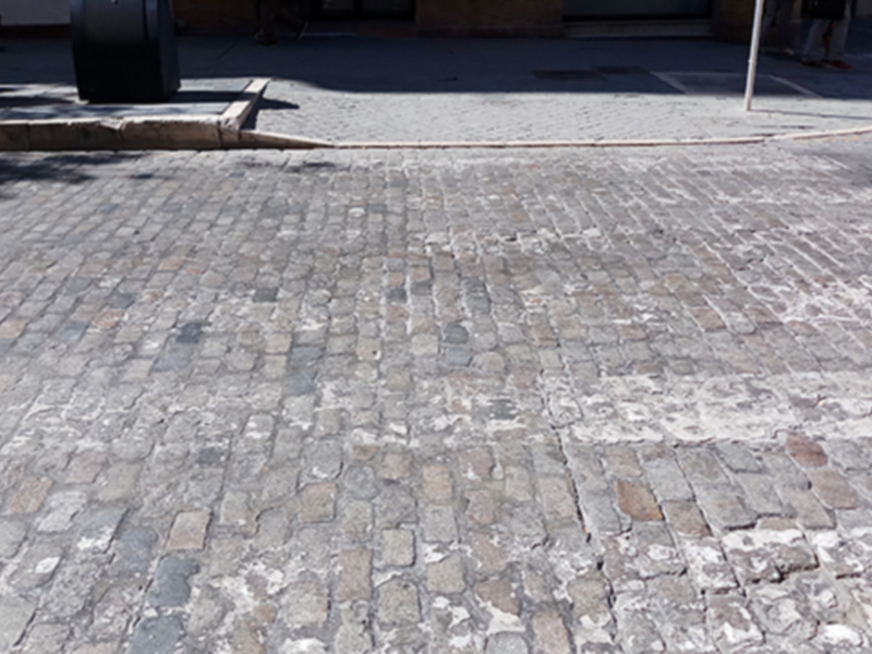 Una calle pavimentada con adoquines de piedra, típica de ciertas zonas históricas o urbanizaciones antiguas. La textura y el color de los adoquines dan un aspecto antiguo y característico a la calle. La imagen también incluye una línea de adoquines que forma un borde para el camino, y en la distancia se pueden ver algunas personas caminando.