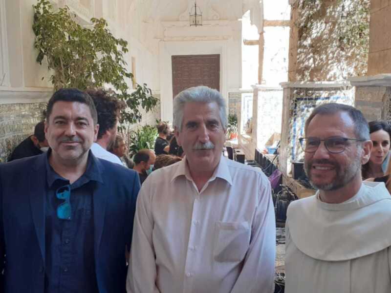 Tres hombres posan frente a una pared de azulejos, uno con un traje oscuro y otro con camisa blanca. El tercero lleva un hábito religioso blanco. La pared está adornada con azulejos de colores y una planta en el fondo.