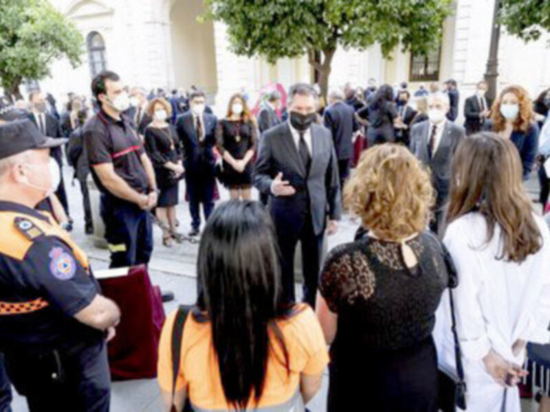 Una multitud de personas, algunos con máscaras, se reúnen en un lugar público. Algunos están hablando y otros escuchando. En el fondo, hay árboles y una estructura arquitectónica clásica. La imagen parece ser de un evento formal o ceremonial.