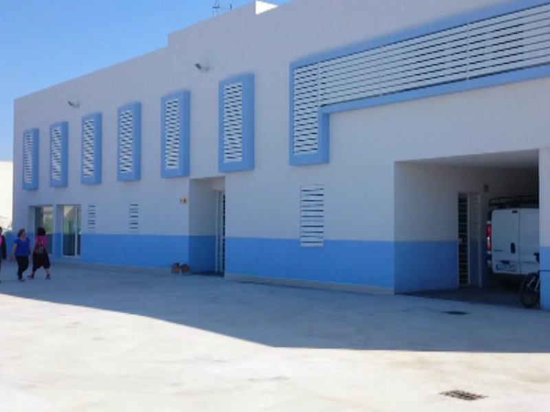 Una fachada de un edificio industrial con ventanas azules y blancas, junto a una entrada principal. En el frente, varias personas caminan hacia la entrada mientras un vehículo blanco está estacionado en el interior. El cielo es claro y azul, indicando buen tiempo.