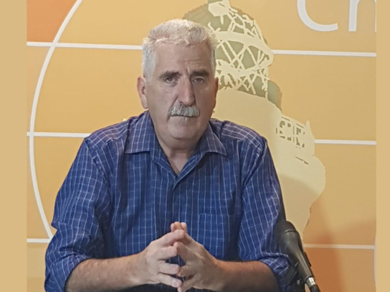 El hombre está sentado frente a un fondo amarillo con una imagen de un globo terráqueo. Está usando una camisa azul a cuadros y tiene las manos juntas en el abdomen.