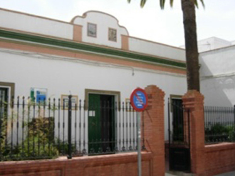 Fachada de una casa blanca con detalles verdes y rojos, rodeada de una verja y una palmera en el jardín.