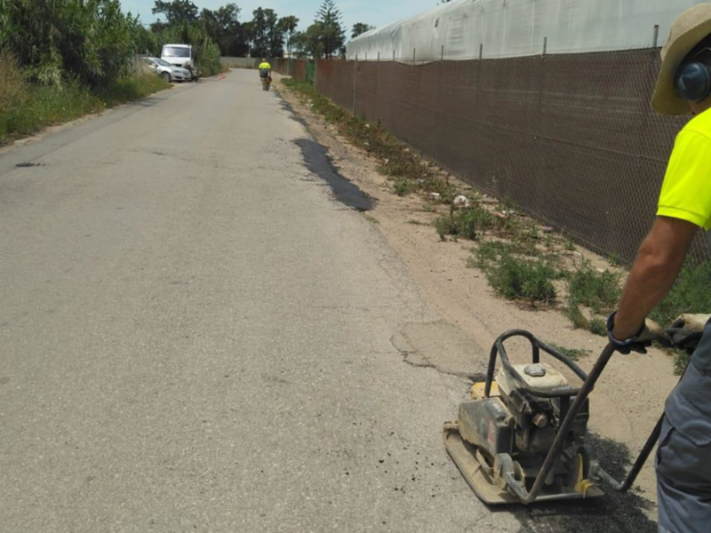 Trabajador utilizando un planchador de asfalto en una carretera secundaria, con vehículos y vegetación en el fondo.