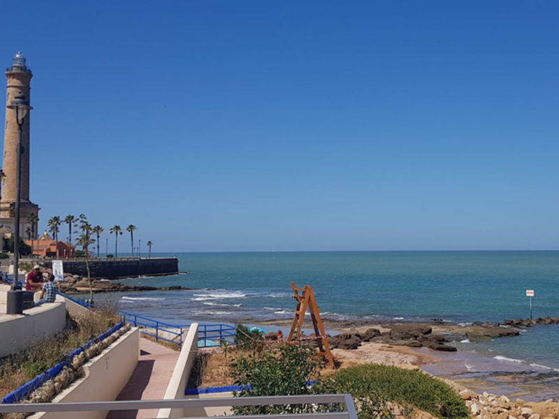 Lighthouse and beach with clear blue sky, rocky shore.