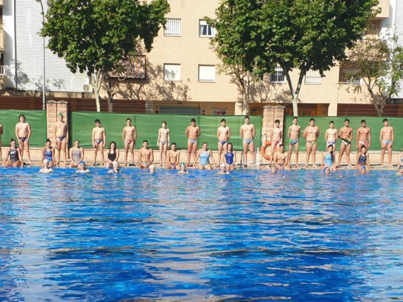 Un grupo de personas en trajes de baño posando en la orilla de una piscina, con árboles y edificios al fondo.