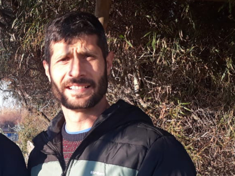 Un hombre con barba y bigote, vistiendo una chaqueta negra y un suéter de punto azul. Fondo natural con vegetación y un árbol en primer plano.