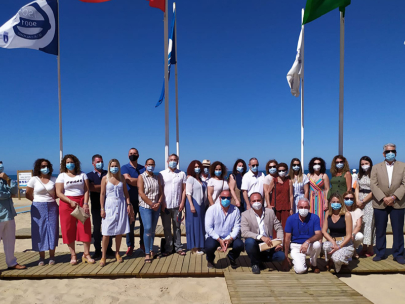 Una imagen de una reunión formal en la playa, con personas vestidas formalmente y banderas en el fondo.