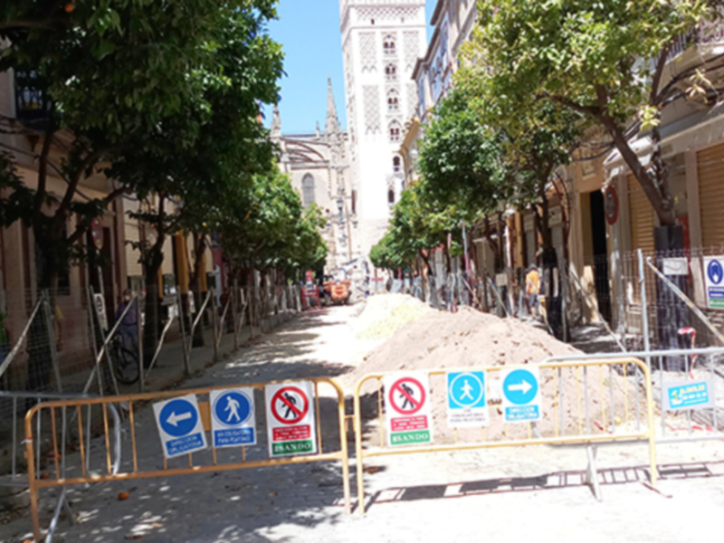 Un camino de hormigón con una pila de arena a la izquierda y varios carteles de construcción en el camino. Hay un edificio con una torre en la distancia y árboles a ambos lados del camino.