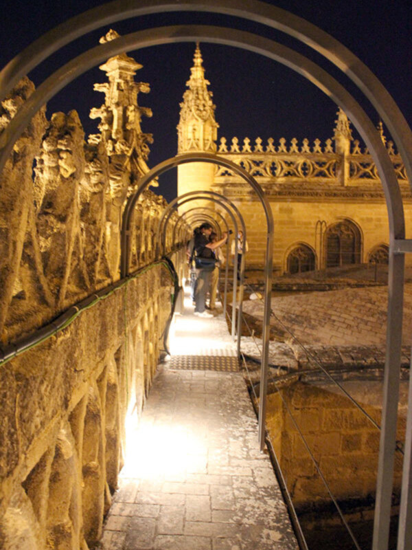 Arco de entrada a un claustro gótico iluminado por la noche, con turistas explorando el lugar.