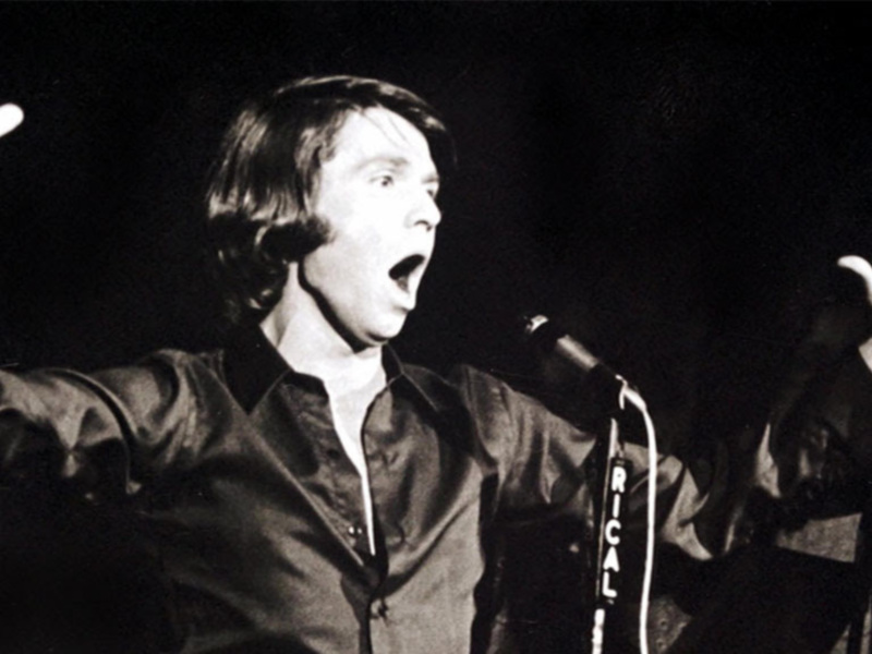 Un hombre con el cabello largo y una camisa negra, con las manos levantadas, parece estar cantando en un escenario oscuro.