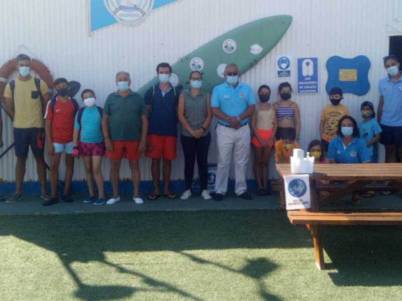 Un grupo de personas y niños posan frente a una pared con un logotipo de club de actividades. La imagen muestra a varios adultos y niños vestidos casualmente, algunos con máscaras faciales. En el fondo se puede ver una pared blanca con un logotipo y carteles, incluyendo uno que dice "Club de Actividades". La escena parece ser en un entorno deportivo o recreativo.