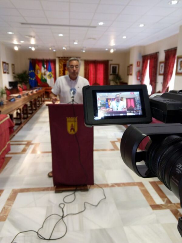 En una sala de conferencias, un hombre se dirige a la audiencia desde un púlpito rojo. En el fondo, banderas ondean y una cámara de video graba la escena. La iluminación es brillante, reflejando en el suelo con patrones geométricos.