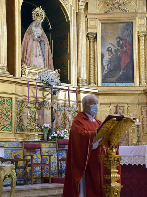 Un sacerdote en una iglesia, vestido con un manto rojo, frente a un altar dorado con una imagen de la Virgen María y un crucifijo. El altar está adornado con flores y tiene un lienzo de pintura religiosa en la pared.