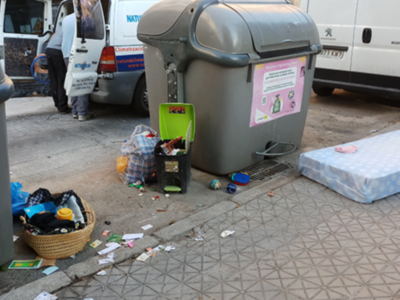 Un contenedor de basura lleno de residuos, incluyendo un pañuelo de papel y una caja de cartón, junto a otros desechos en el suelo. Un cartel informativo está pegado al contenedor, y una camioneta blanca con el logo de Peugeot está estacionada en el fondo.