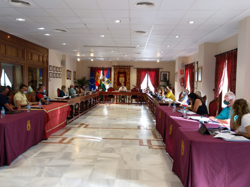 Sala de reuniones con mesas rojas, personas sentadas, cortinas coloridas y ventanas.
