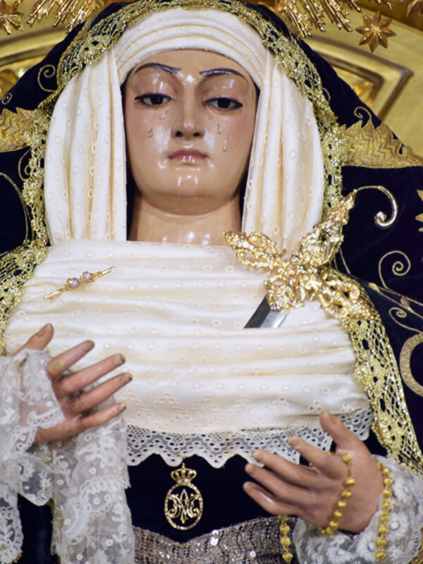 Una estatua de una Virgen María vestida con un manto blanco y dorado, con detalles en oro y piedras preciosas. La Virgen tiene una expresión serena y está sosteniendo un objeto en su mano izquierda. La estatua es parte de una procesión religiosa, con un fondo dorado y estrellas.