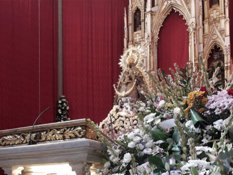 Una estatua religiosa en un altar adornado con flores y una profunda decoración de oro. El fondo es rojo, y la estatua está sobre un pedestal con detalles dorados.
